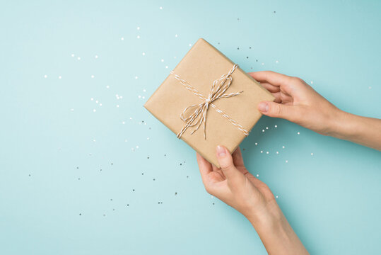 First person top view photo of hands giving craft paper gift box with twine bow over shiny sequins on isolated pastel blue background with copyspace