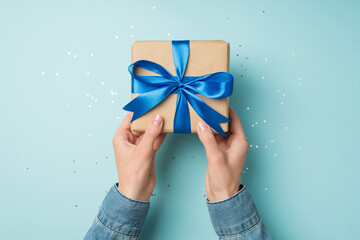 First person top view photo of hands in denim shirt holding craft paper giftbox with vivid blue...