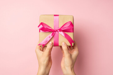 First person top view photo of hands giving stylish craft paper giftbox with pink satin ribbon bow on isolated pastel pink background