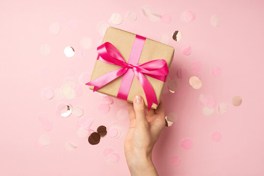 First person top view photo of girl's hand holding stylish craft paper giftbox with pink satin ribbon bow over shiny confetti on isolated pastel pink background