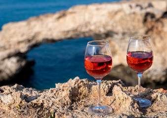 Two glasses of rose dry white wine served on rocks in blue sea bay with Love Bridge on background near Ayia Napa touristic town on Cyprus