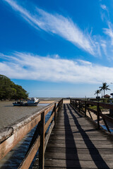 Fototapeta premium View of the beach of Rio das Ostras with the meeting of the river in Rio de Janeiro. Sunny day, blue sky. Yellow sand and some rocks. Wooden bridge to cross. Boats and fishing on the pier.