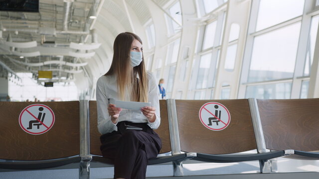 Confident business woman in a mask sitting at the airport, holding a passport and plane tickets, keeps a distance in a pandemic. Waiting at the airport. Travel concept. Woman looking at tickets