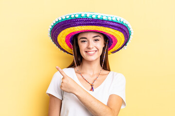 young hispanic woman smiling cheerfully, feeling happy and pointing to the side. mexican hat concept - obrazy, fototapety, plakaty