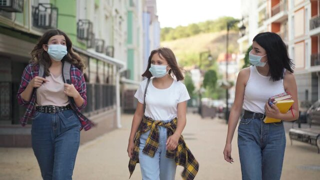Portrait of three Caucasian college students in Covid face masks strolling on city street with backpacks talking. Confident teenage girls walking outdoors after classes on coronavirus pandemic