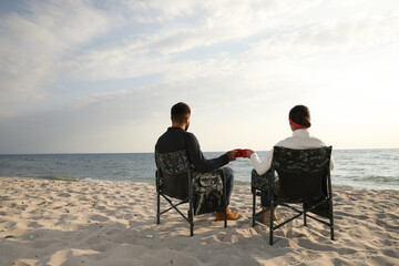 Couple sitting in camping chairs and clinking mugs on beach, back view