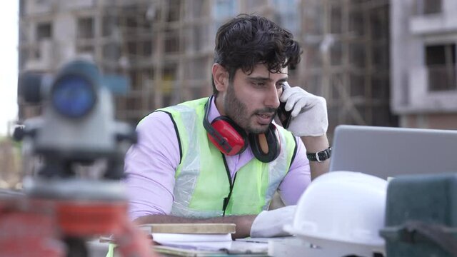 Indian civil engineer talking on smartphone while working on laptop at construction site.Worker wearing earmuff at construction site outdoor.Asian male wearing safety jacket and talking on mobile.

