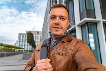 Selfie portrait of smiling handsome millennial man with backpack on one shoulder standing at building background on city street. Successful man just got new business opportunity.