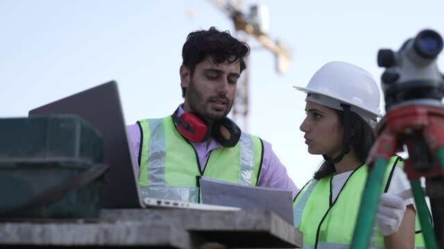 Two Indian engineers on Construction Site using papers and Looking at laptop Plans while working on under construction site.Young Asian man and woman using theodolite and laptop at construction site.