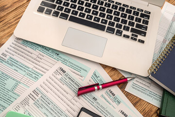 on a wooden table are a laptop, calculator, dollar bills, pen and tax forms
