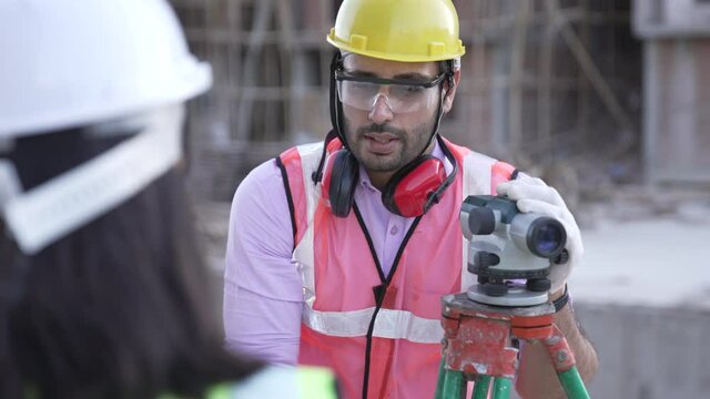 Two Indian man and woman engineers surveyor take measures with theodolite use papers talking together stand helmet hardhat geodesy building digital engineering real estate teamwork infrastructure.