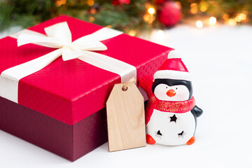 A toy Santa penguin, a red gift box with a white ribbon and a bow, a wooden blank form for greeting text on a background of a branch of a fir tree with balls. Christmas and New Year's decorations