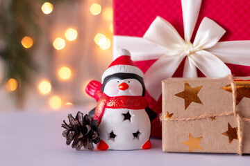 A toy penguin in a Santa's hat near a gift boxes, cone on a background of a fir tree and blurred bokeh lights of garland. Christmas and New Year's atmosphere