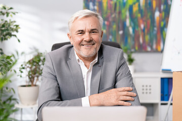 Close-up of face of smiling man in shirt jacket, guy with gray hair stubble in old age is sitting on comfortable chair in office of company is looking into camera friendly boss hands crossed on chest