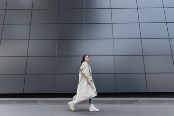 trendy woman in long raincoat and white boots walking along grey wall