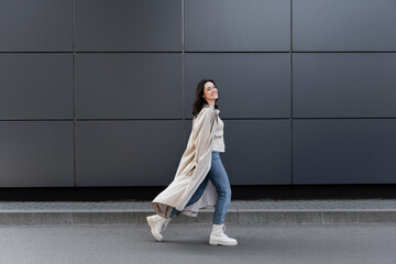 Fototapeta na wymiar cheerful woman walking along grey wall in long coat and white leather boots