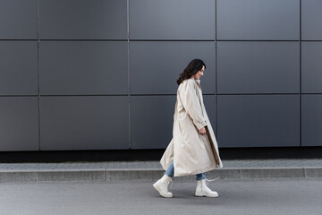 side view of young woman in white boots and long raincoat walking near grey wall