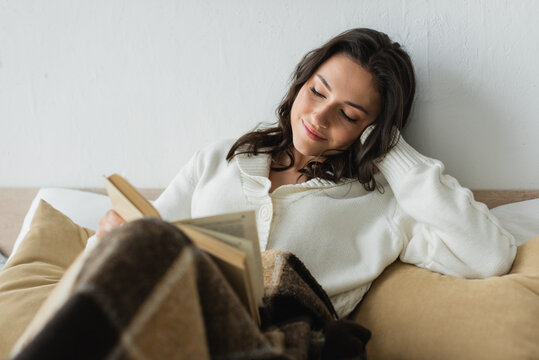 Smiling Woman In White Cardigan Reading Book In Bed Under Checkered Blanket