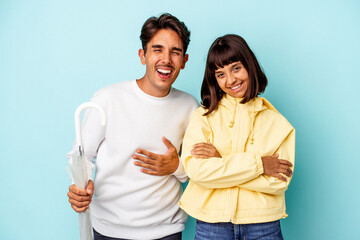 Young mixed race couple holding umbrella isolated on blue background laughing and having fun.