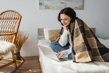 smiling woman using computer on bed while sitting under checkered blanket
