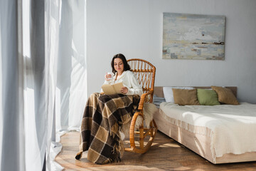 brunette woman reading book in bedroom while sitting in rocking chair under plaid blanket - obrazy, fototapety, plakaty