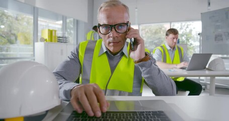 Mature architect typing on computer and consulting client on cellphone in office