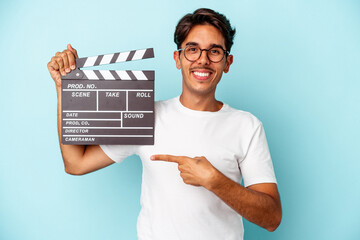 Young mixed race man holding clapperboard isolated on blue background smiling and pointing aside,...