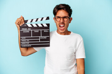 Young mixed race man holding clapperboard isolated on blue background screaming very angry and...