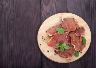 Fototapeta premium Pieces of jerky meat with basil leaves lie on a wooden cutting board on a wooden background.