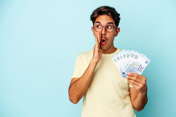 Young mixed race man holding bills isolated on blue background is saying a secret hot braking news and looking aside