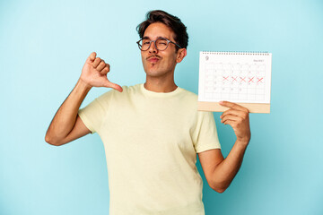 Young mixed race man holding calendar isolated on blue background feels proud and self confident,...