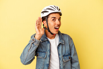 Young mixed race man wearing a helmet bike isolated on yellow background trying to listening a gossip.