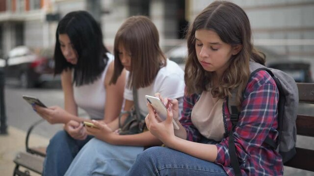 Bored Caucasian teenage girl yawning sitting on city street with college groupmates surfing social media on smartphones. Portrait of disinterested device addicted students outdoors on bench
