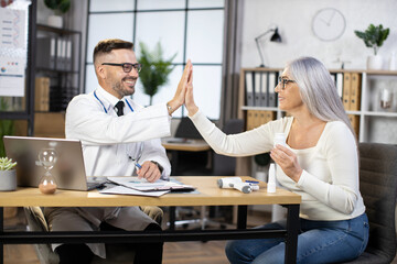 Happy senior woman giving high five to male doctor while sitting at office of medical service. Successful recovery of aged patient. Competent health treatment and medical service.