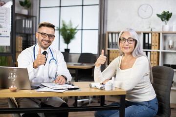 Caucasian bearded doctor in white lab coat and eyeglasses sitting at desk with senior woman,...