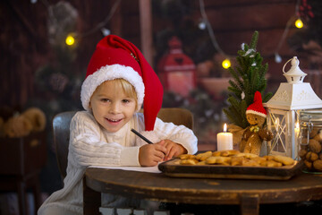 Cute toddler child, boy on Christmas, writing letter to Santa Claus and eating cookies at home, wooden cottage