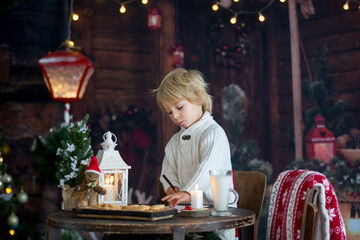 Beautiful toddler child, blond boy, writing letter to Santa Claus and eating cookies at home, cozy atmosphere