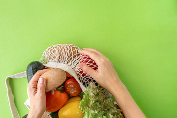 top view of female hands reaching for organic eco vegetables from mesh shopping bag on green background. Caring for the environment and avoiding the plastic concept.