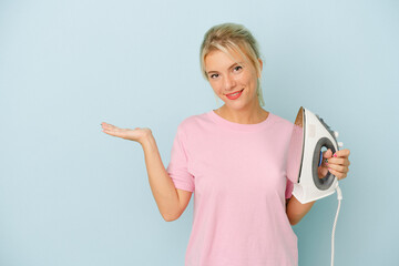 Young Russian woman holding iron isolated on blue background showing a copy space on a palm and...