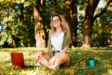 Smiling young woman freelancer meditates, relaxes during remote work outdoors. Teenage girl with glasses sits in lotus position with laptop and disposable cup of coffee on green lawn in park outdoors