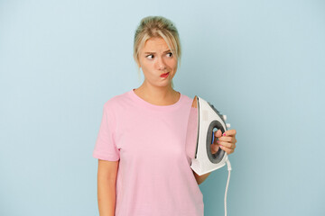 Young Russian woman holding iron isolated on blue background confused, feels doubtful and unsure.