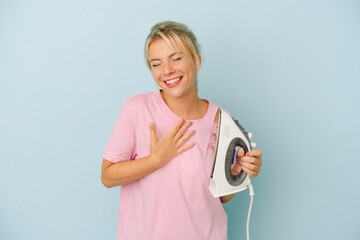 Young Russian woman holding iron isolated on blue background laughs out loudly keeping hand on chest.