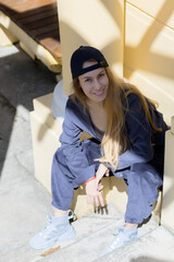 Smiling long-haired blonde woman dressed in sports style sits near a light plaster wall in park architecture