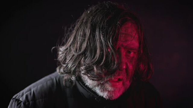 A scary portrait of a creepy and dirty homeless man. The man says something and looks at the camera. The terrible picture in dark and red tones is creepy. Fear overwhelms me.