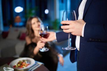 Beautiful young couple in elegant wear spending evening time at home for romantic dinner. Man giving glass of red wine to charming woman. Close up.