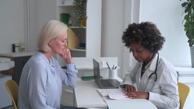 Female doctor talking to patient and smiling, sitting at table during working day in clinic spbas. Young medical expert discusses symptoms of disease with old lady and looks with smile, gives advice
