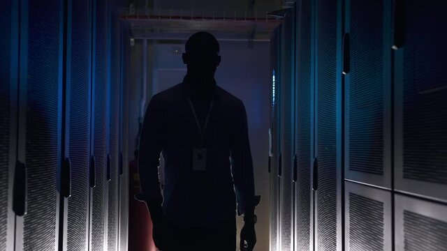 Male engineer walking through dark interior during working day at data center spbas. Front view of young African American man walks and poses in room with server equipment. Closeup view of experienced