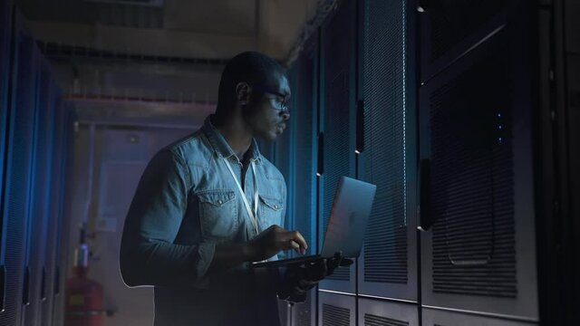 Male engineer using laptop and providing network support during working day in server room spbas. Closeup view of young American man types text and looks at computer screen, does job and examines at