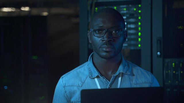 Front of young businessman using computer while sitting at table on farm render on dark night spbas. Close-up view of American African man looks at laptop screen then poses for camera with serious