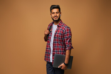Smiling indian man in casual close with laptop and backpack on pastel background